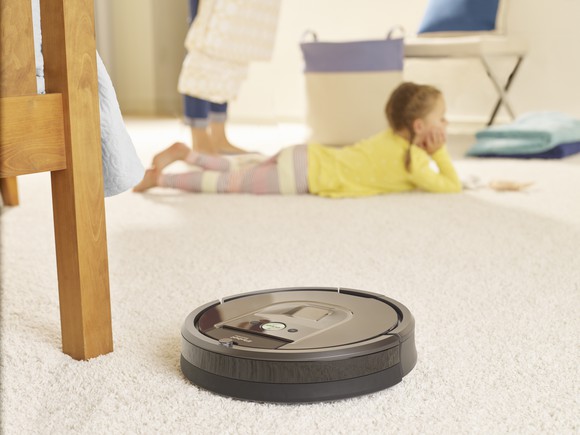 iRobot Roomba 980 cleaning carpet on a carpet next to a wooden chair, with a young girl in the background.