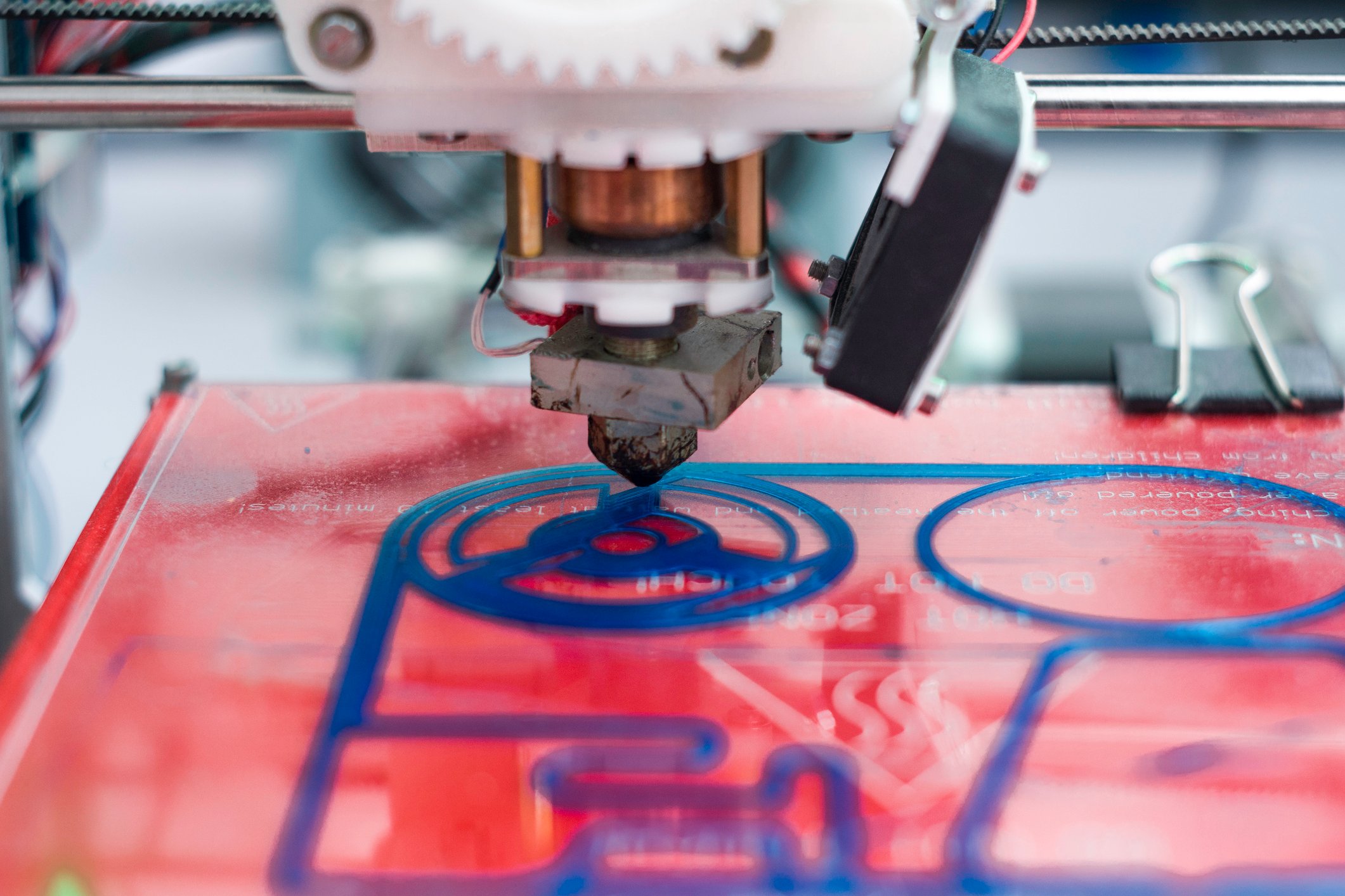 Close-up of a 3D printer head just starting to deposit a blue polymer resin on a red work surface. 