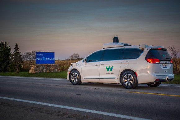 A white Chrysler minivan with Waymo markings and visible self-driving sensor hardware is shown passing a sign reading "Welcome to Pure Michigan".