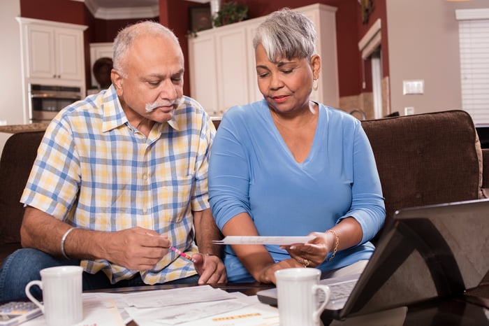 An older couple sits side by side in a living-room setting. They look at a piece of paper as they sit in front of a table covered in other papers and holding two drink mugs.