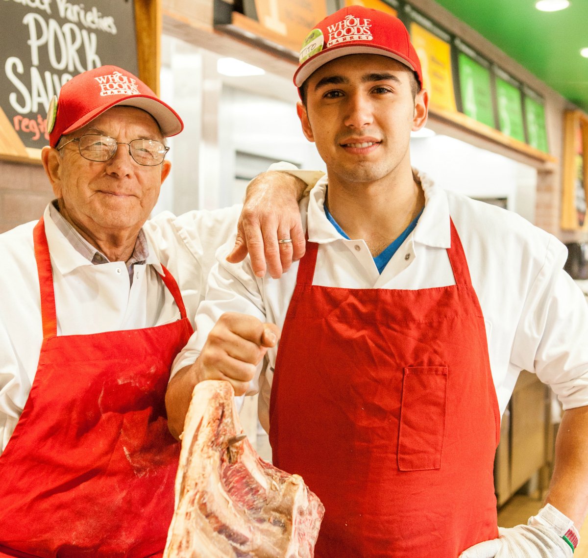 Two workers wearing red Whole Foods hats and red aprons stand together at a Whole Foods. 
