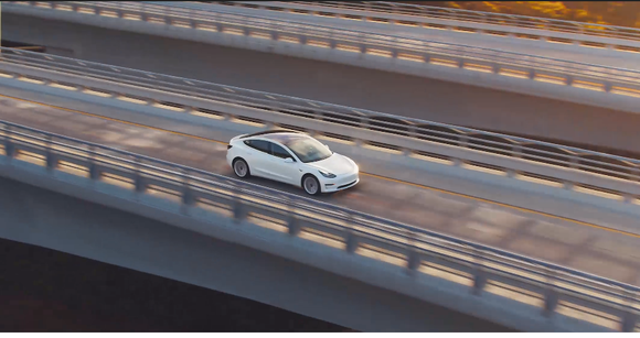 A view from above and somewhat to the side of a white Model 3 driving across a long and high bridge.