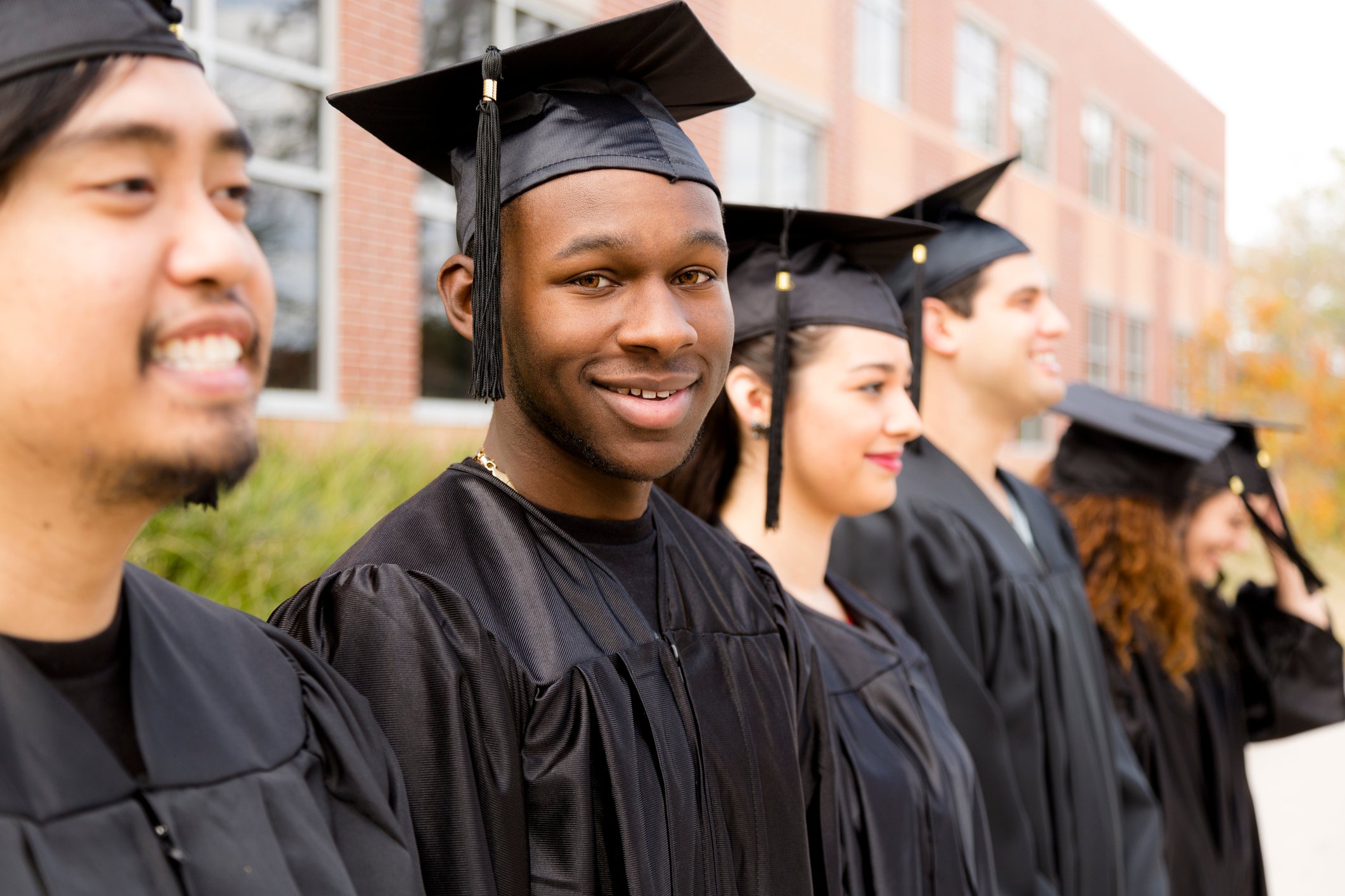 College students in a row at a graduation ceremony wearing black robes in front of a school building.