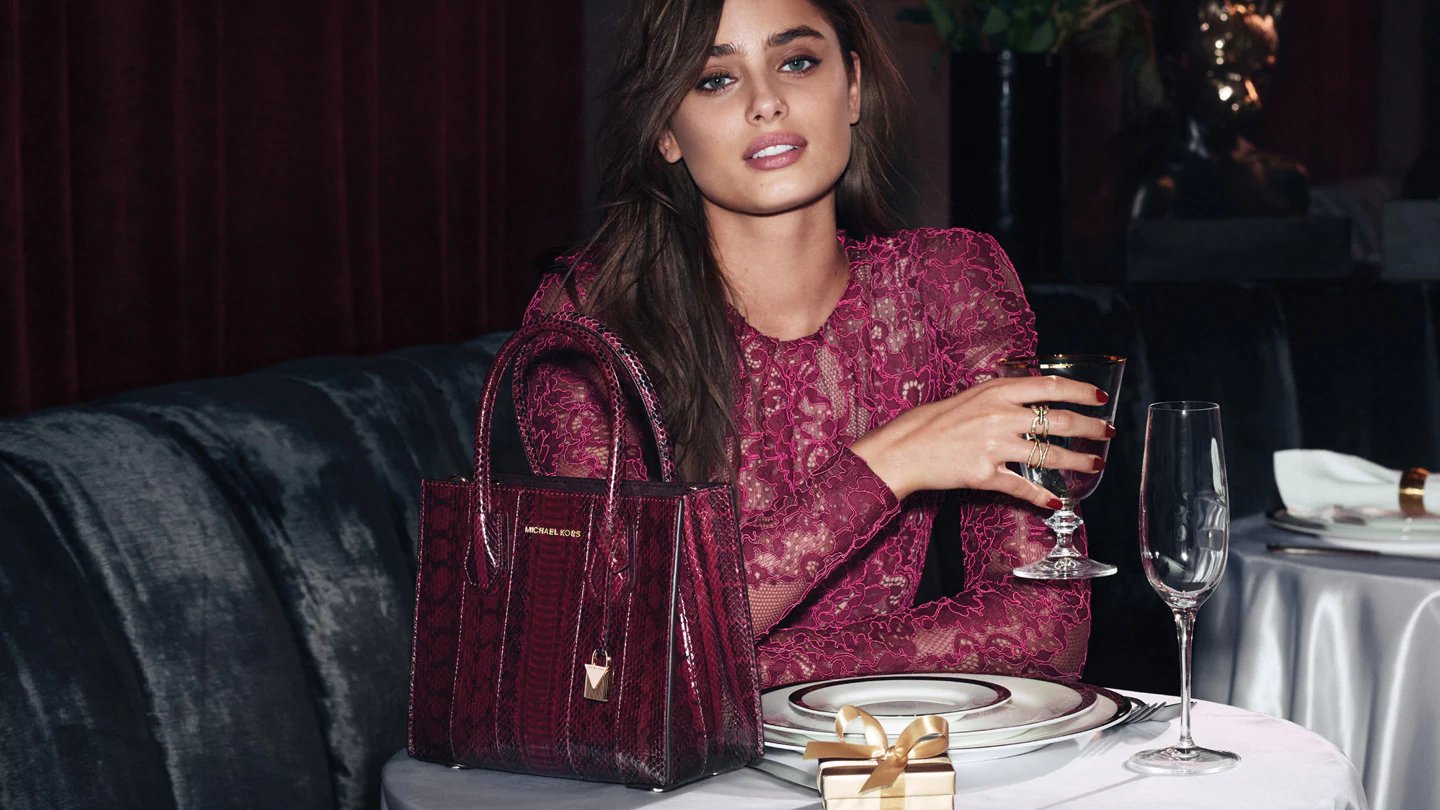 A model sits at a restaurant table with a glass and a Michael Kors bag