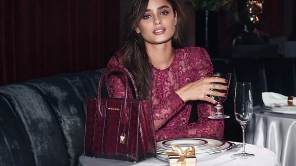 A model sits at a restaurant table with a glass and a Michael Kors bag