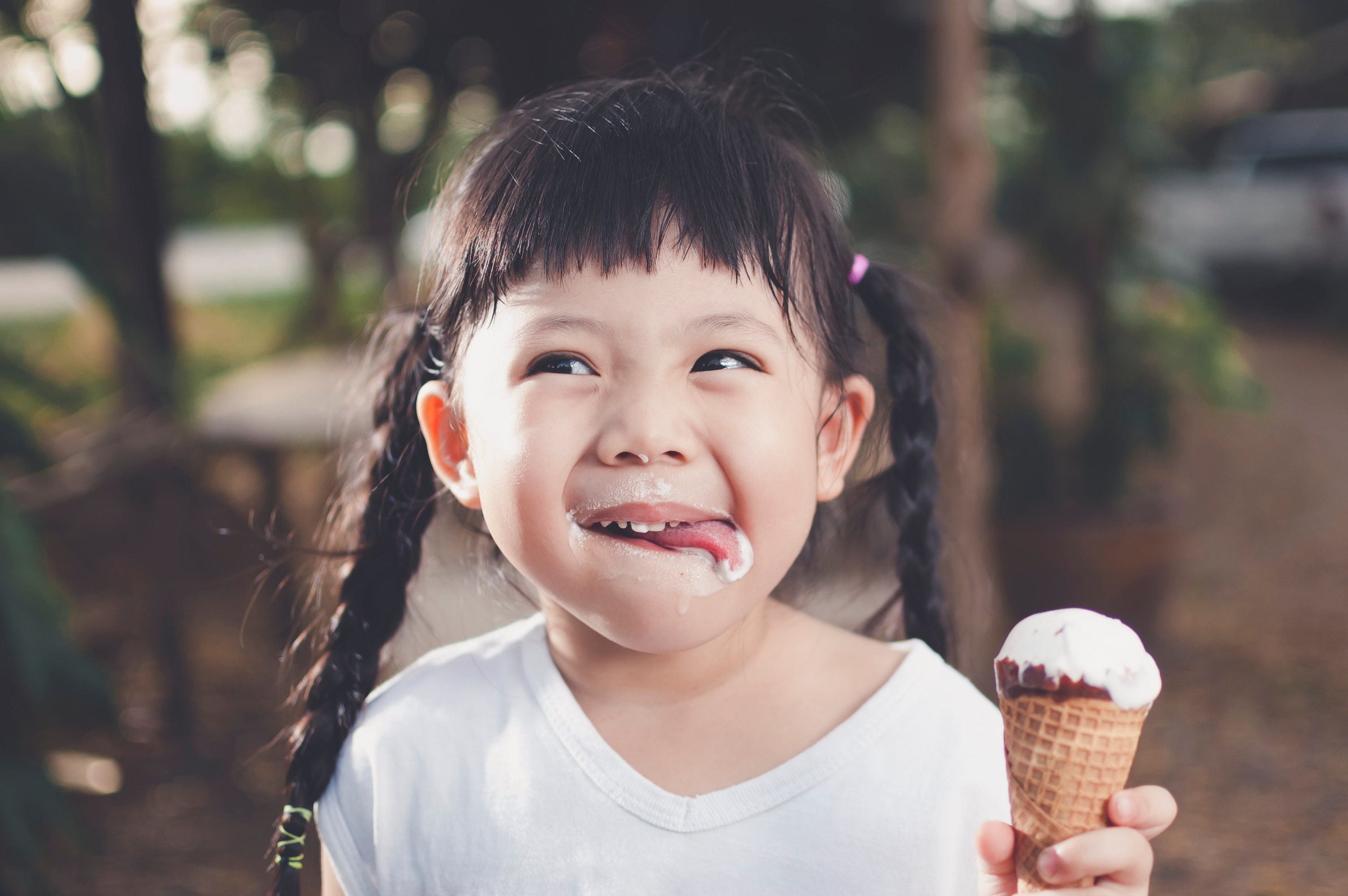 A young, smiling girl in pigtails holds an ice cream cone and smiles as she licks ice cream off her face. 