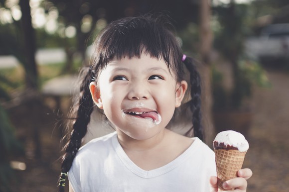 A young, smiling girl in pigtails holds an ice cream cone and smiles as she licks ice cream off her face. 