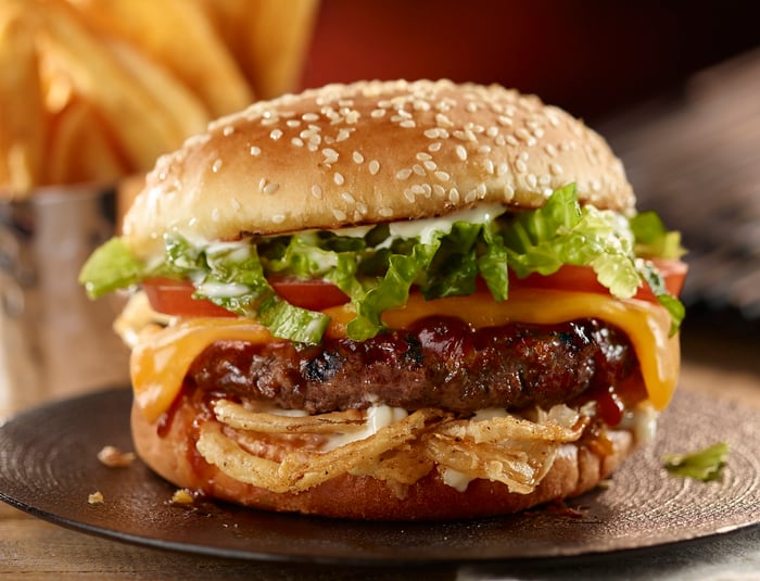 Burger with sesame seed bun, lettuce, tomato, cheese, onions, and BBQ sauce on a plate.