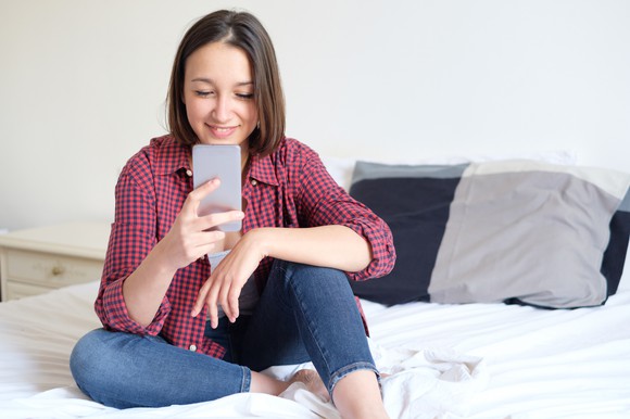 Young woman sitting on bed looking at smartphone