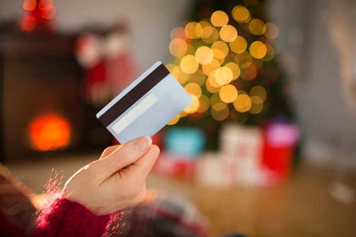 A shopper holds a credit card while looking at presents around a Christmas tree.