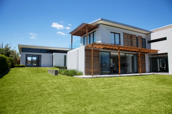 White two-story modern home with white walls and wood accents, and a green grass lawn.