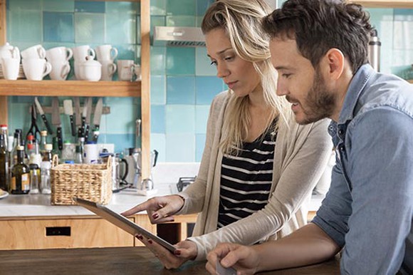 Man and woman in kitchen (cups and bottles on a shelf in background) looking at a tablet and discussing something.