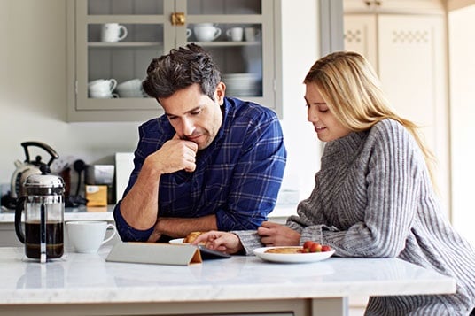 Couple sitting at table peering at tablet, some berries and a coffee maker in front of them.