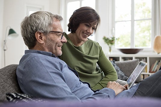 Couple sitting on couch staring at tablet, grinning. Window behind them in picture.