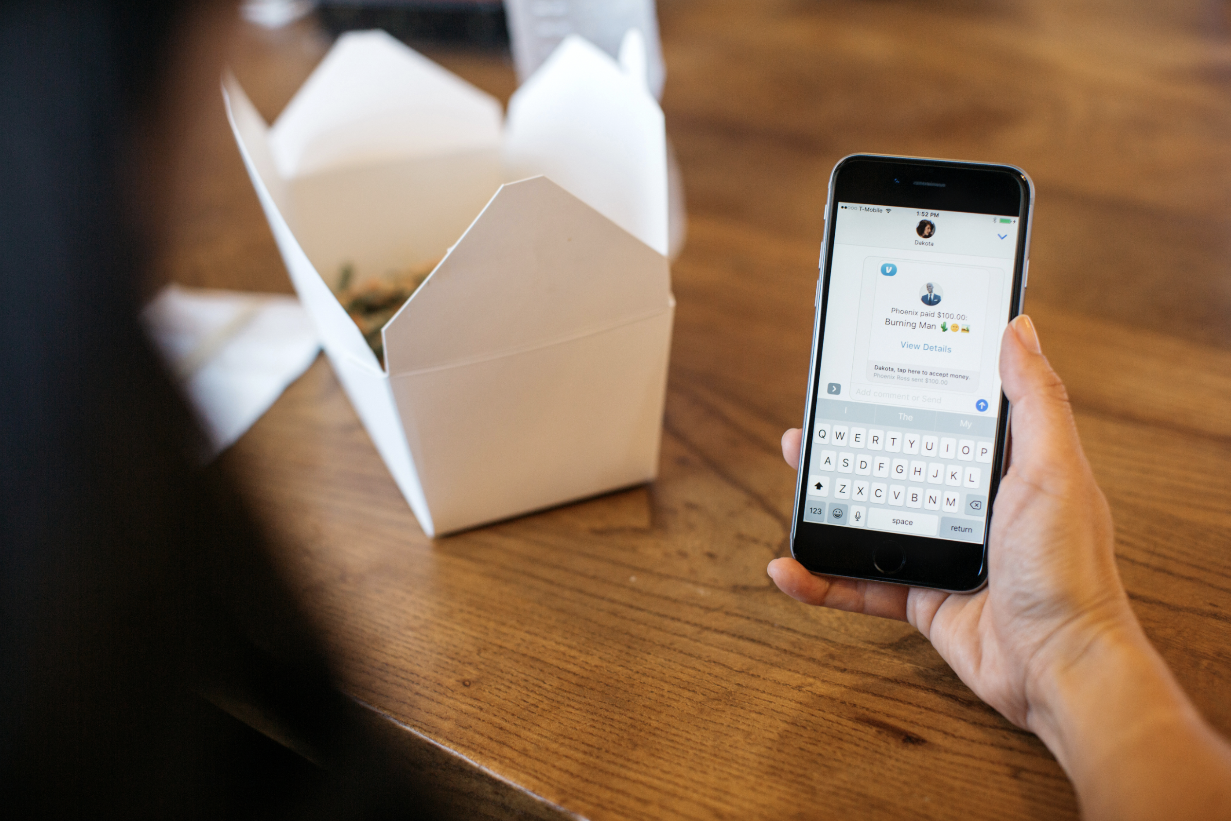 A table with food and woman's hand holding a smartphone with the Venmo app displayed.