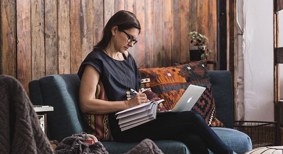 Female entreprenuer working at home on couch with laptop and paper notebook.