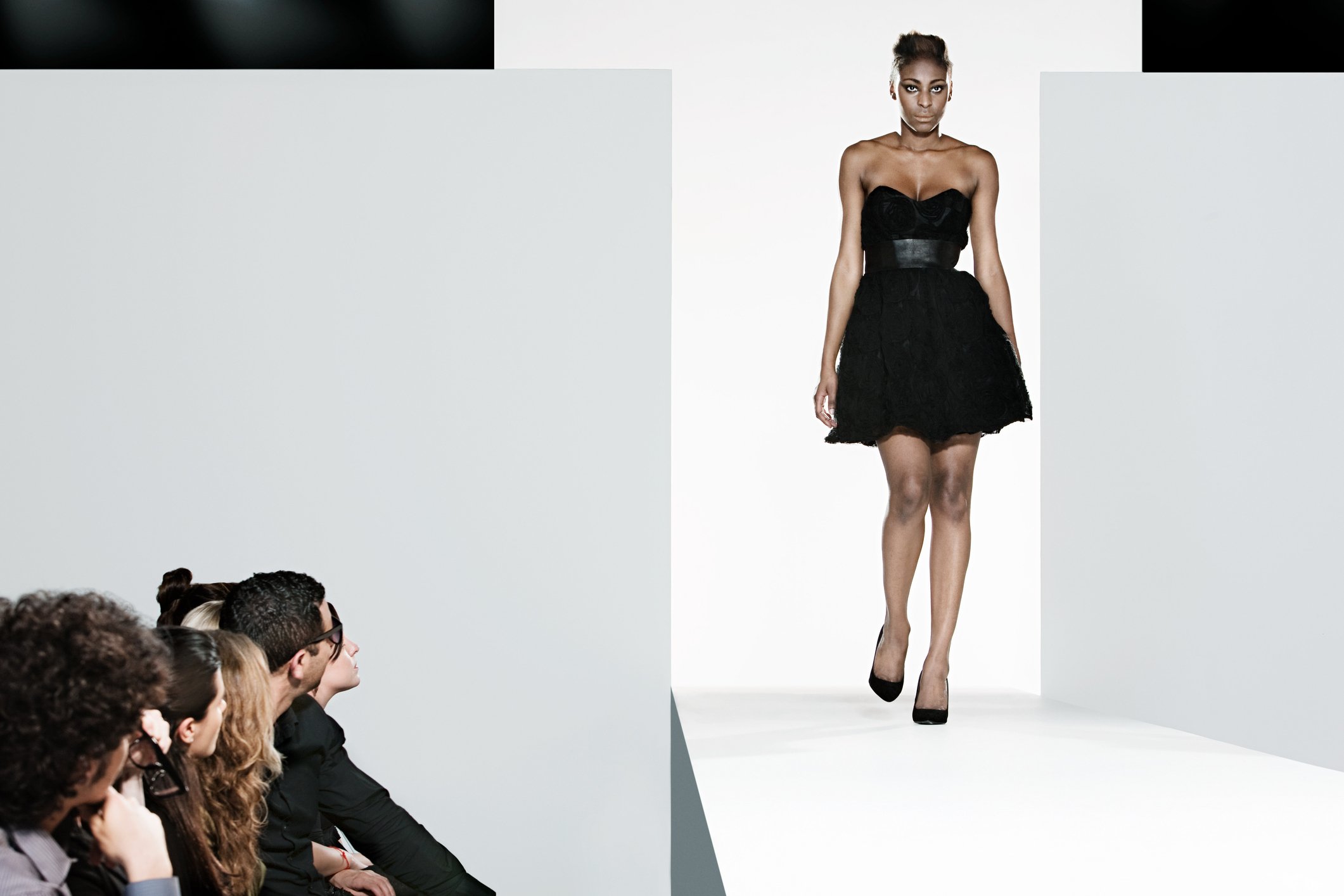 Model in low-cut, short, tight black dress with flouncy skirt on fashion show runway