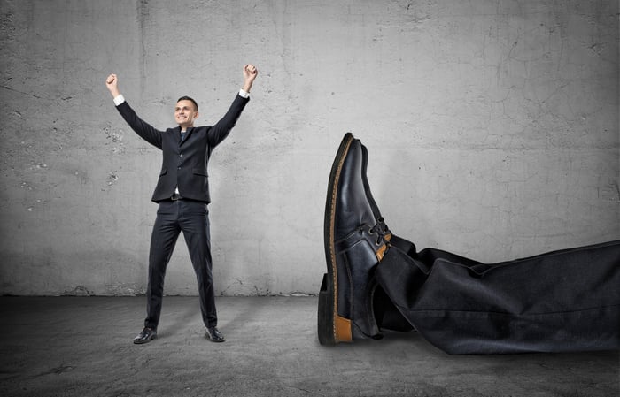 Small man with hands raised next to feet of giant man lying on ground