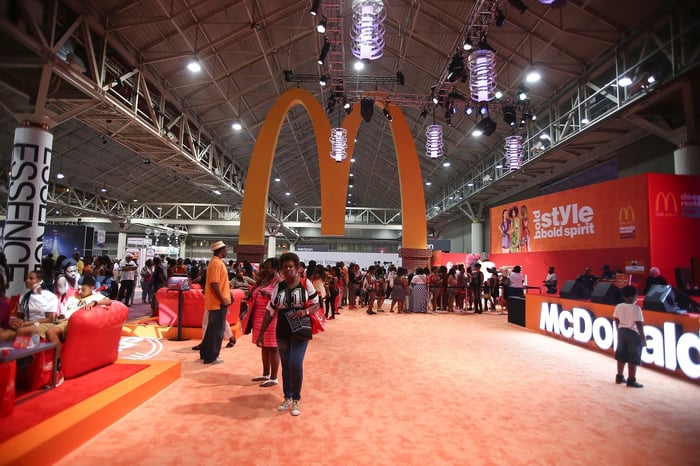 Golden Arches hanging from the ceiling of a convention hall, with a booth nearby with the McDonald's logo and many people in attendance.