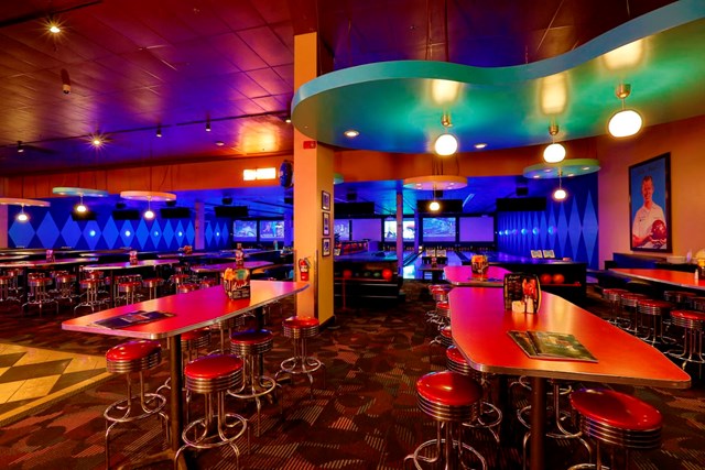 The interior of a Dave & Buster's, with a bowling alley in the background and bar stools and tables in the foreground.