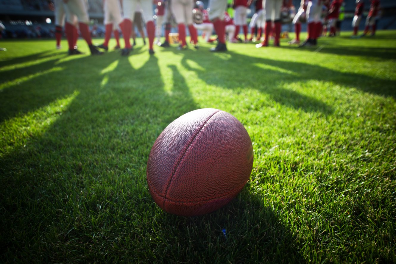 A football on a football field, with players in the background.