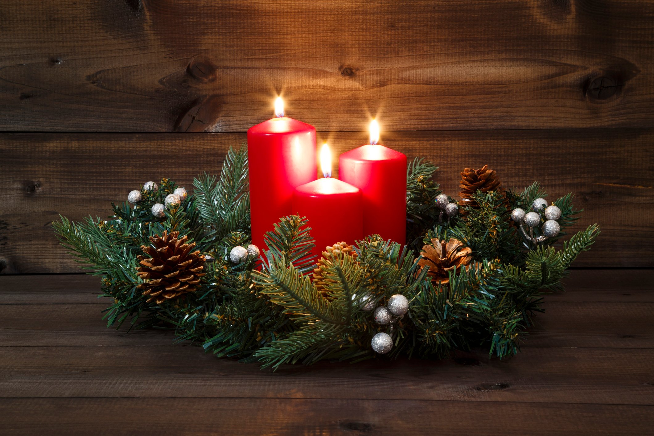 Three red candles lit in the center of a wreath of evergreen branches and pine cones.