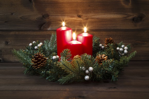 Three red candles lit in the center of a wreath of evergreen branches and pine cones.