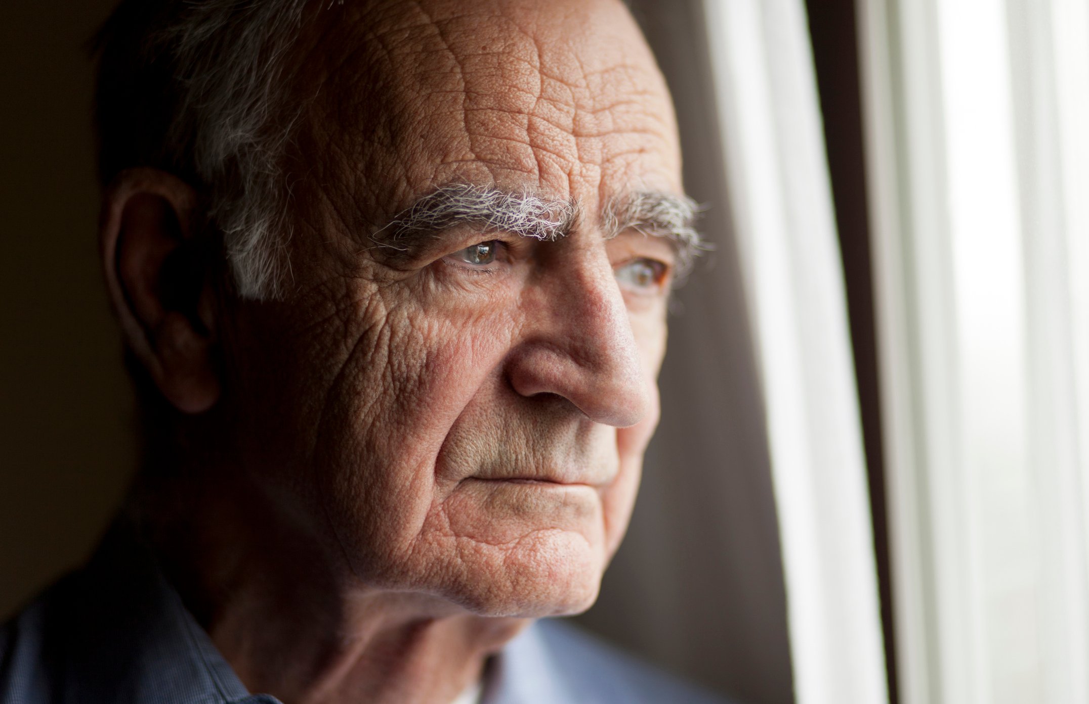 A worried elderly man staring out of a window in his home. 
