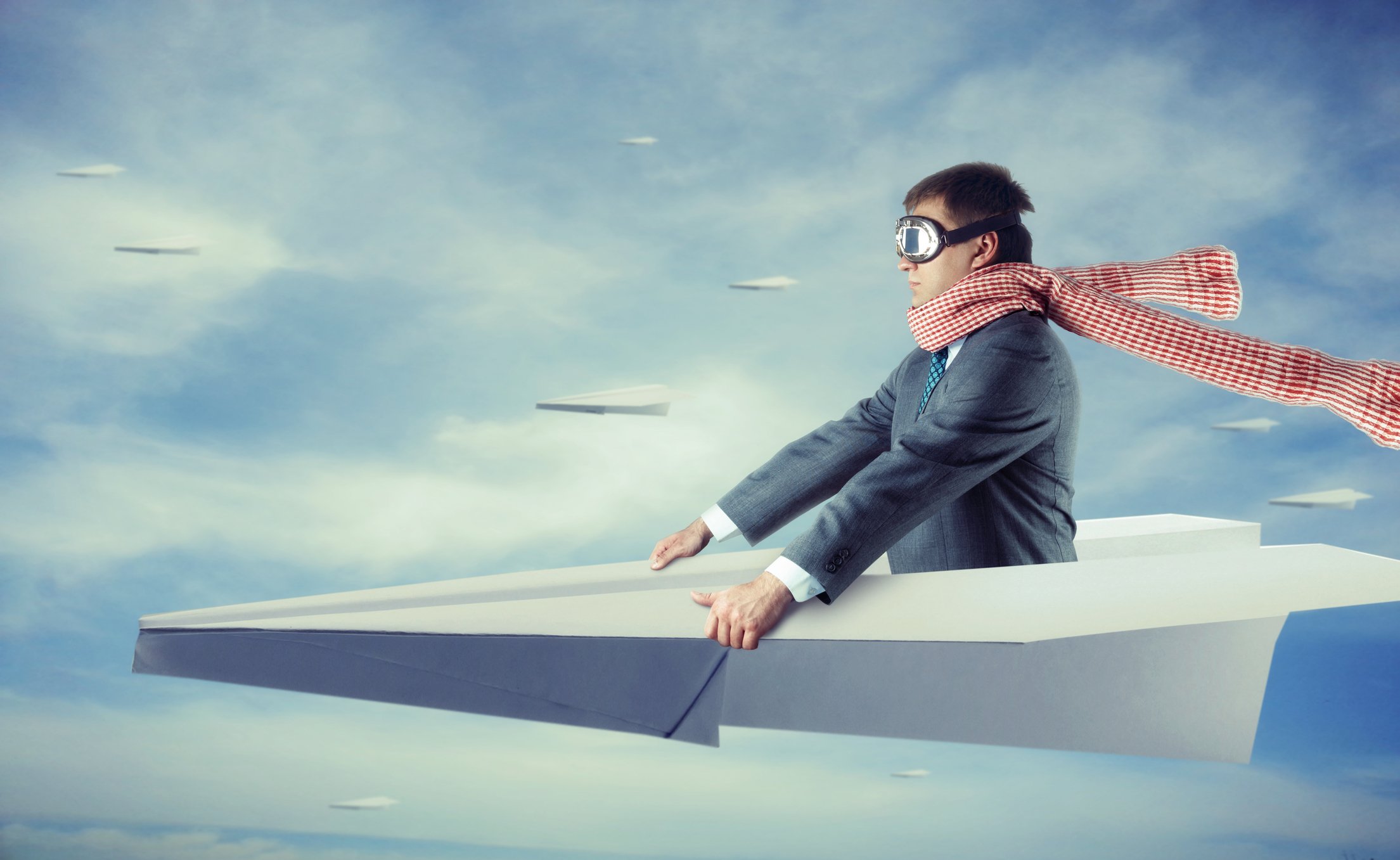 A man in a gray suit, goggles, and a long scarf rides a paper airplane in the sky