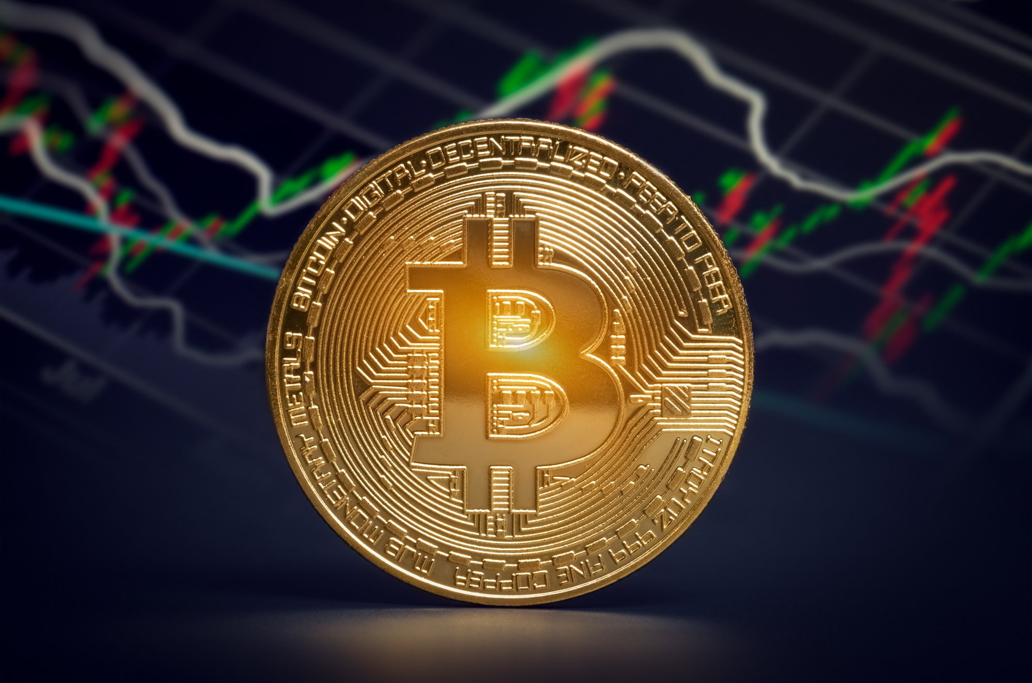 Gold coin with a bitcoin symbol on it in front of a stock chart.