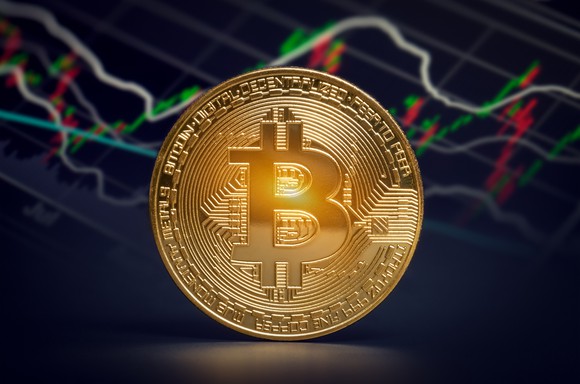Gold coin with a bitcoin symbol on it in front of a stock chart.