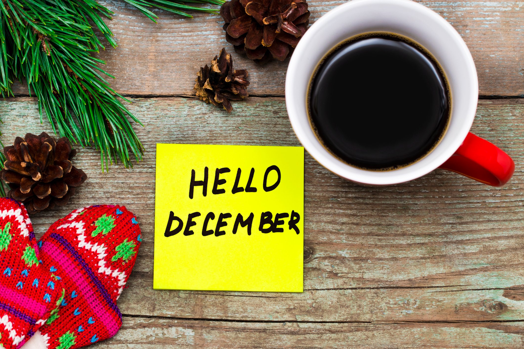 Post-it note that says Hello December on wood surface, surrounded by coffee cup, glove, and pinecones. 