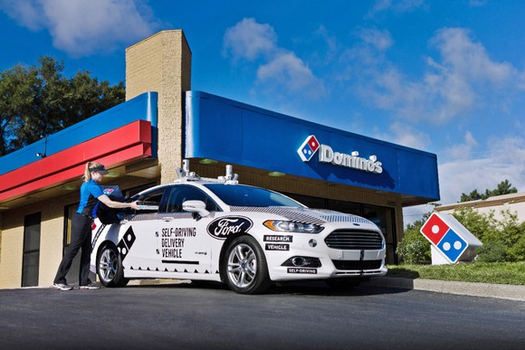 A white Ford Fusion Hybrid with self-driving sensor hardware and Ford and Domino's Pizza logos is shown parked outside a Domino's restaurant.
