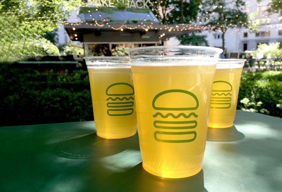 Three drinks in Shake Shack cups on a green table, with a Shake Shack restaurant in the background.