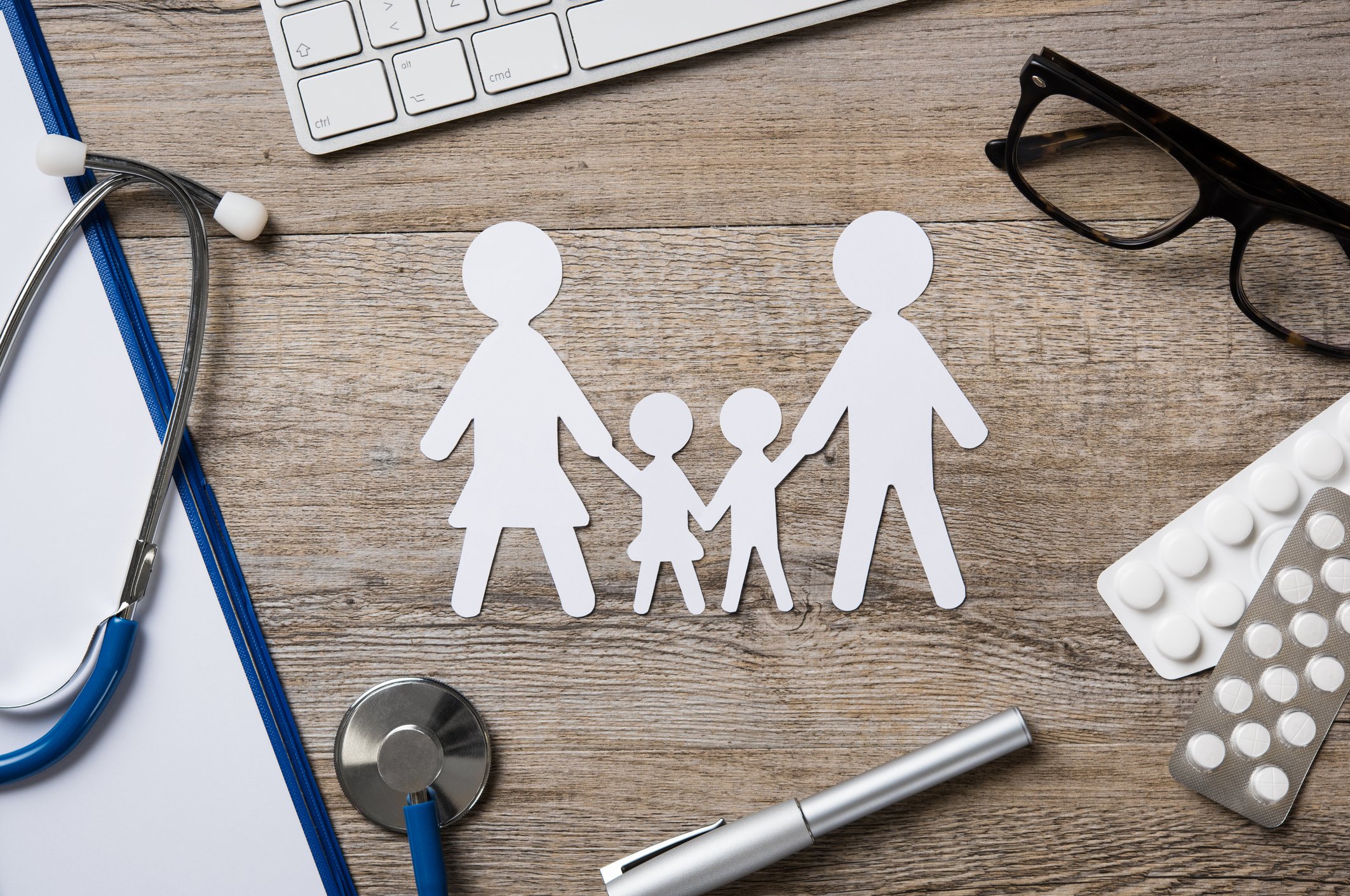 A paper cut out family rests on a desk beside a stethoscope, pen, glasses, and prescription tablets.