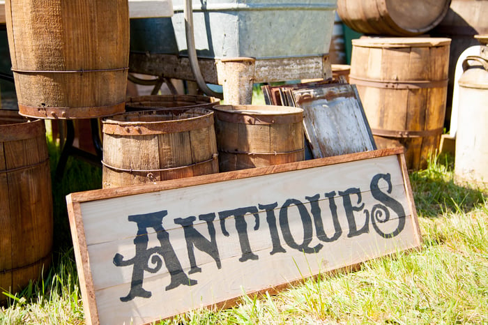 Sign with the word antiques on it, resting outdoors on the grass against a pile of wooden barrels.