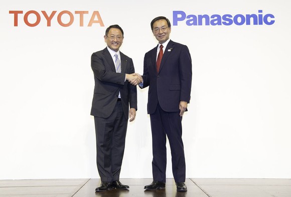 Akio Toyoda and Kazuhiro Tsuga are standing, shaking hands, in front of a backdrop with Toyota and Panasonic corporate logos.