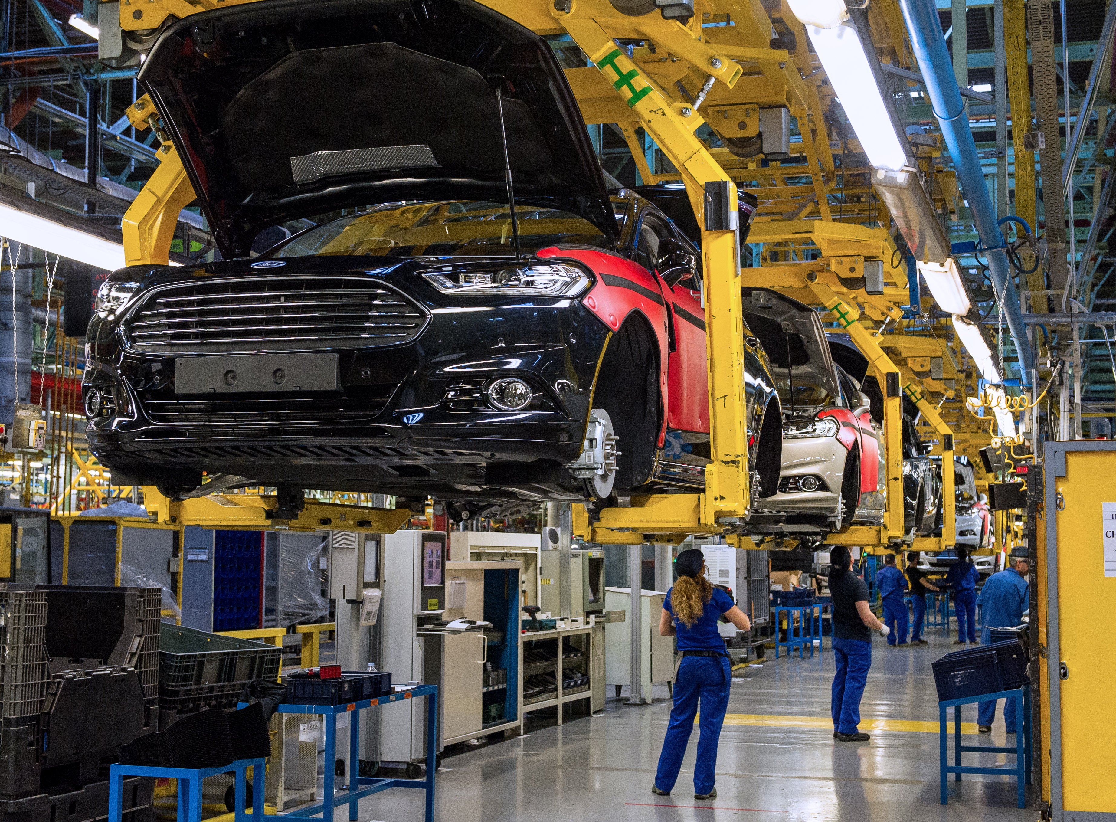 Partially-completed Mondeo sedans are shown in cradles, several feet above the factory floor, with workers underneath. 