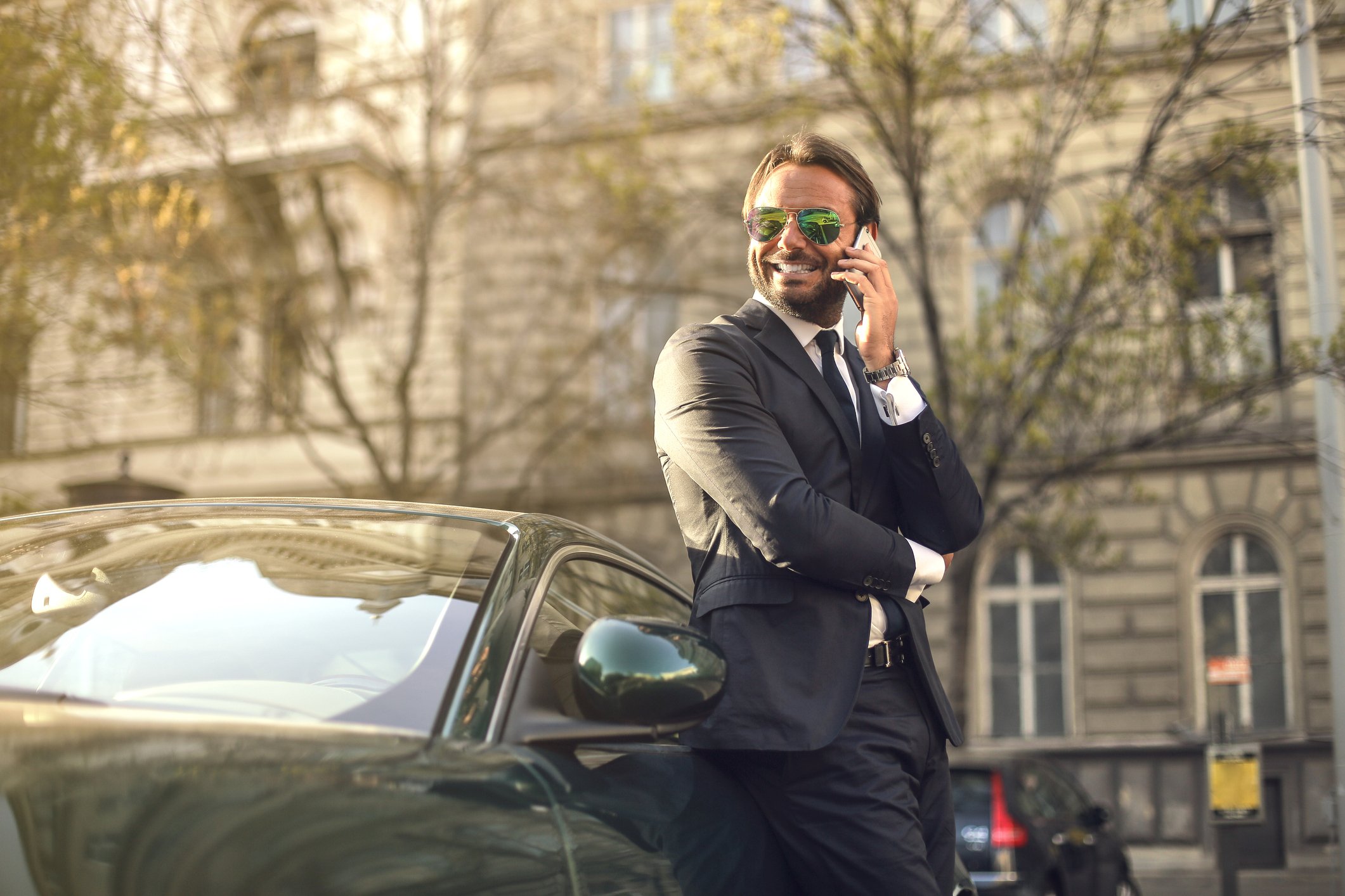 Man in a suit talks on a phone in front of a luxury car.