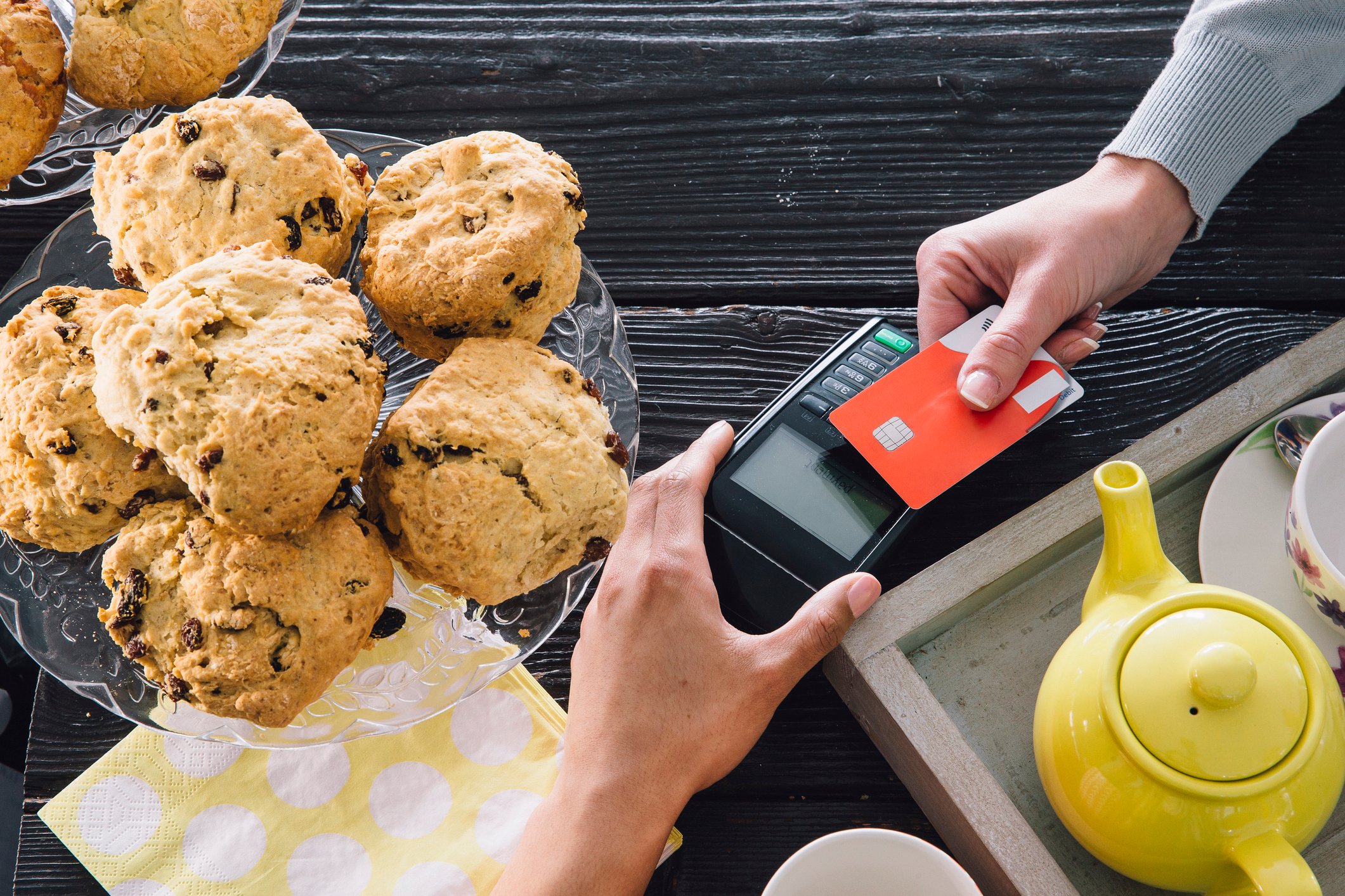 A man's hand holds credit card against contactless credit card reader over a table with a platter of muffins. 