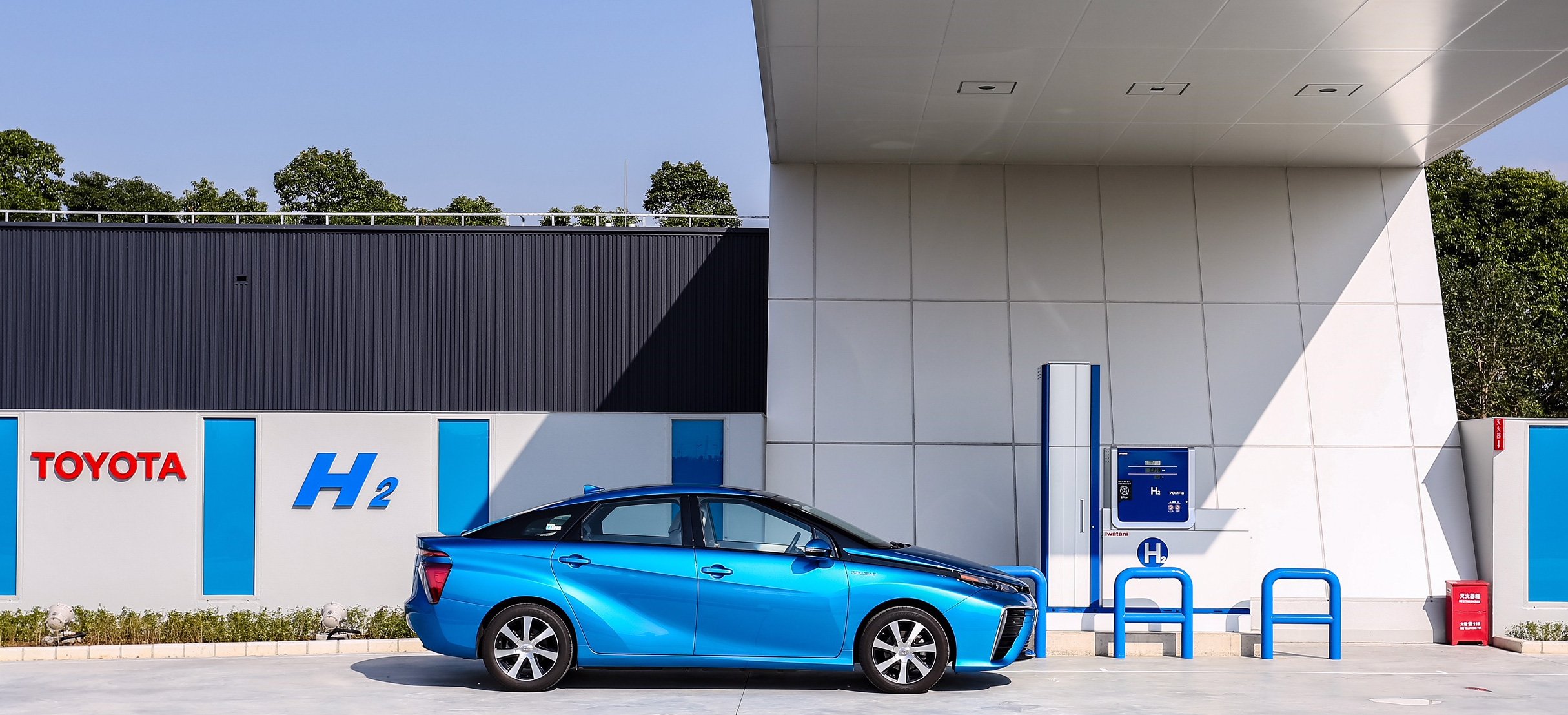 A blue Toyota Mirai fuel-cell sedan parked at a refueling station. Toyota H2 is printed on a wall behind.