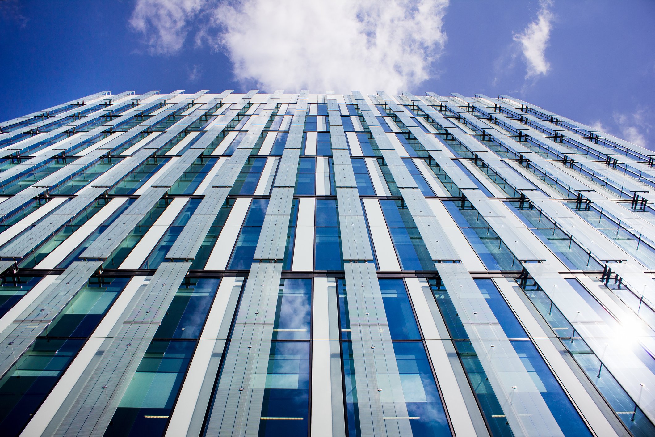 The surface of a glass and metal building with a bright but cloud-filled blue sky above it 