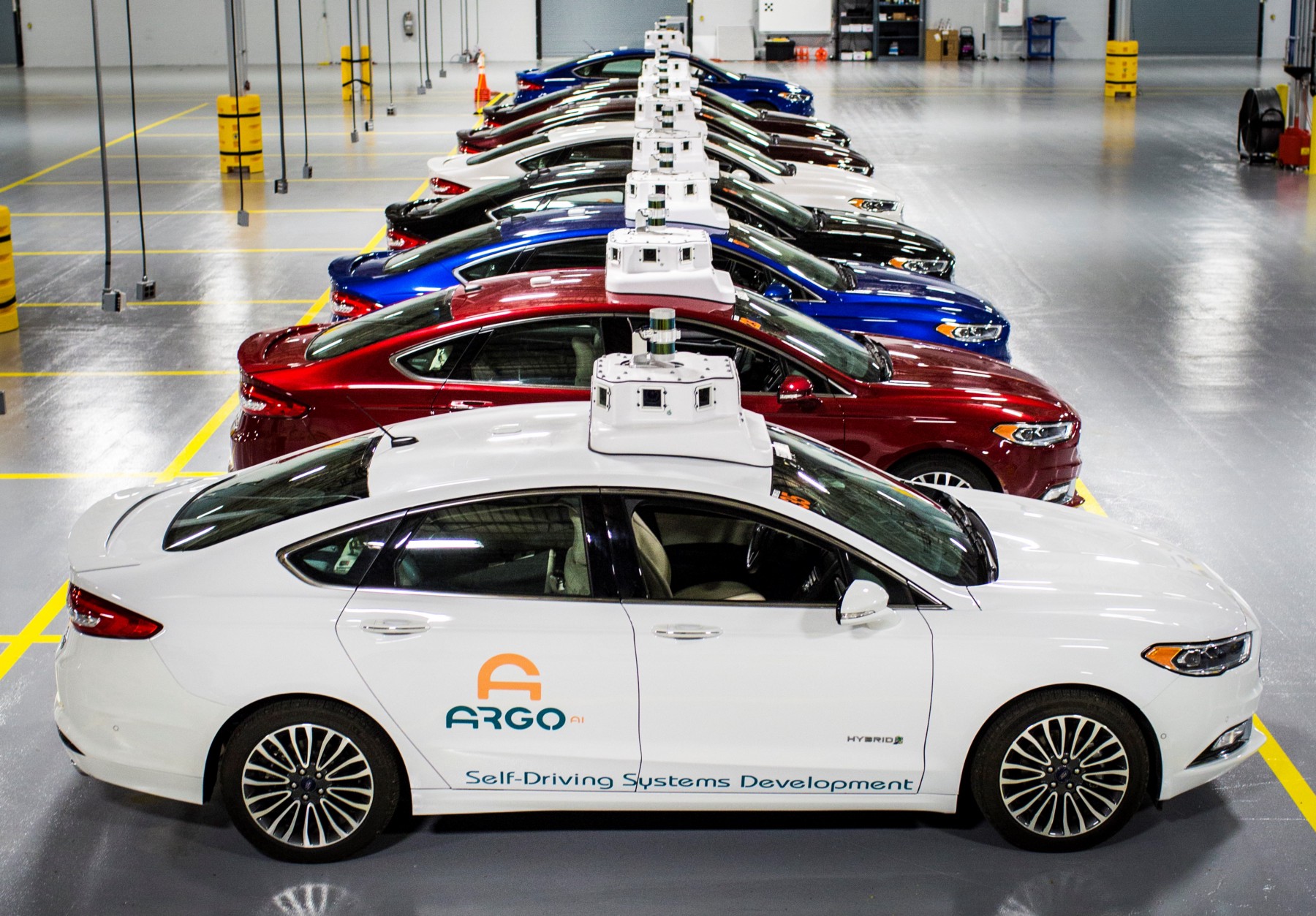 A row of Ford Fusion Hybrid sedans with Argo AI logos and visible self-driving sensor hardware, parked in a garage. 