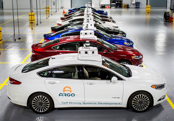 A row of Ford Fusion Hybrid sedans with Argo AI logos and visible self-driving sensor hardware, parked in a garage. 