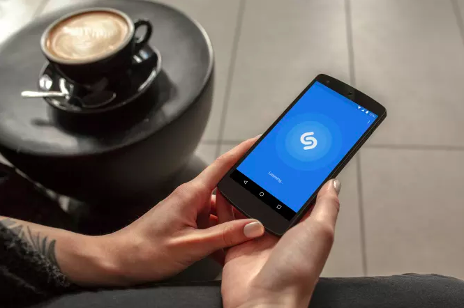 Hands hold an Android phone with Shazam pulled up and a coffee table with a latte stands nearby