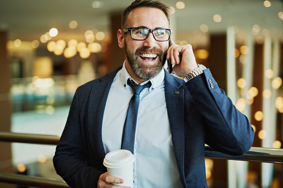 A smiling man in jacket and tie speaks on the phone, while holding a covered white beverage cup in the other hand.