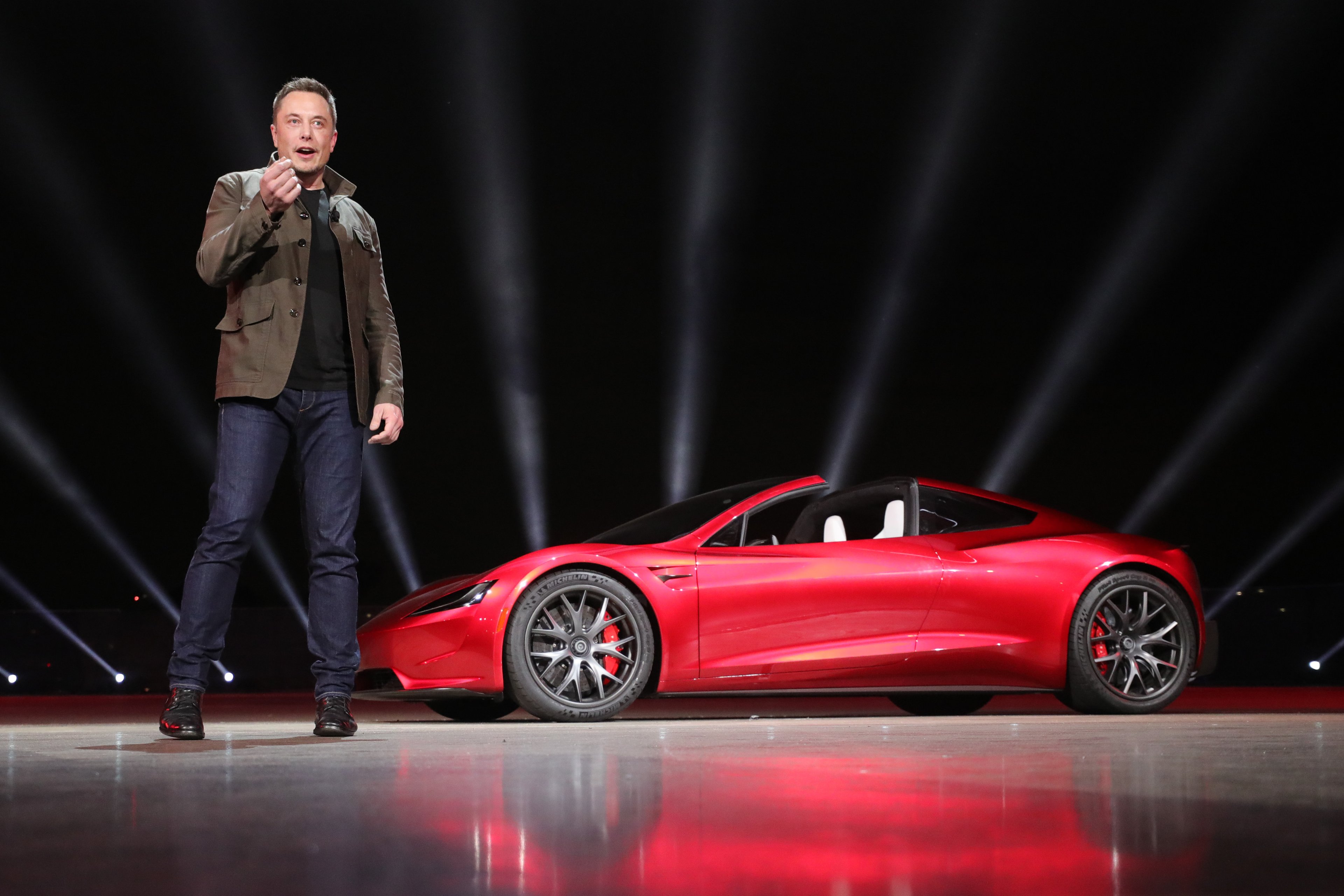 Elon Musk speaking on a stage next to a red second-generation Tesla Roadster. 