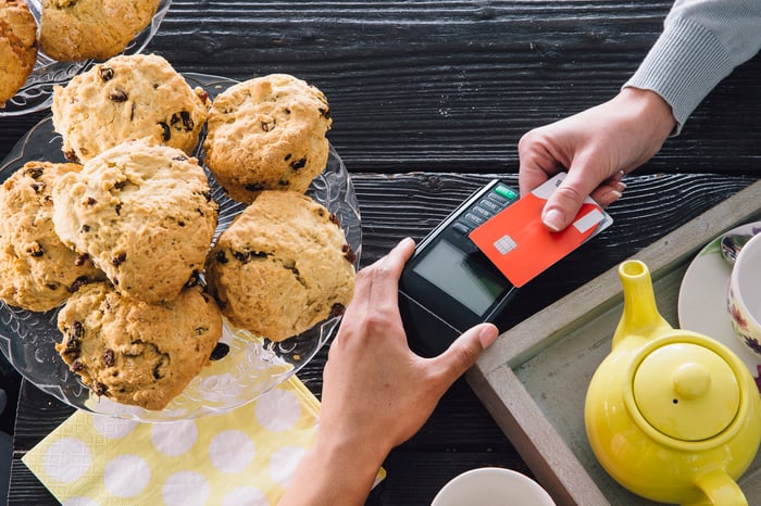A person paying for an item at a bakery counter by holding a chip credit card over a card reader terminal. 