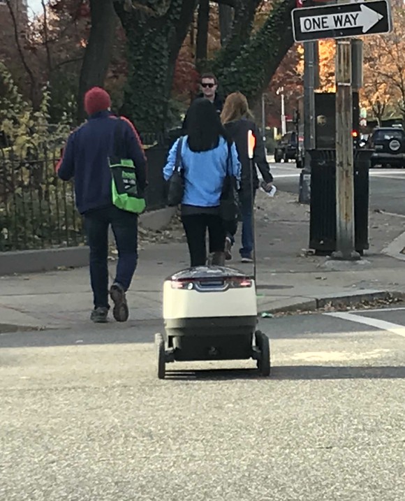 Small white robot on two wheels crossing the street and trailing three people