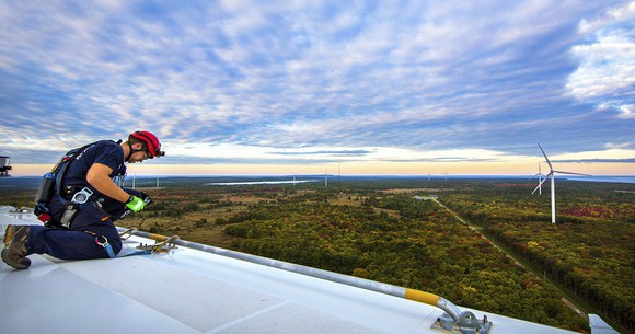 Worker on top of a wind turbine blade, with other wind turbines in a lightly forested area on a partly cloudy day.